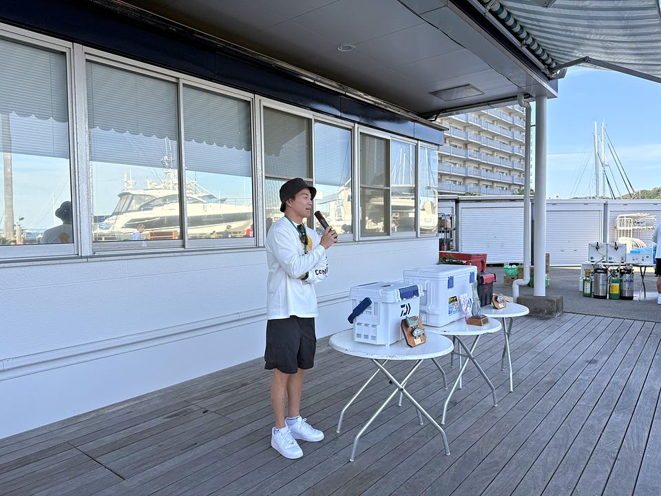 クラブハウス前のデッキでマイクを握る BOL全国幹事長の杉本氏と、賞品が並ぶテーブル