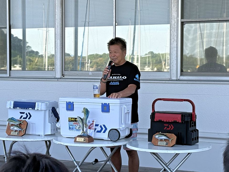 トロフィーと賞品が並ぶテーブルの後ろでマイクを握る MAHALO の谷本キャプテン