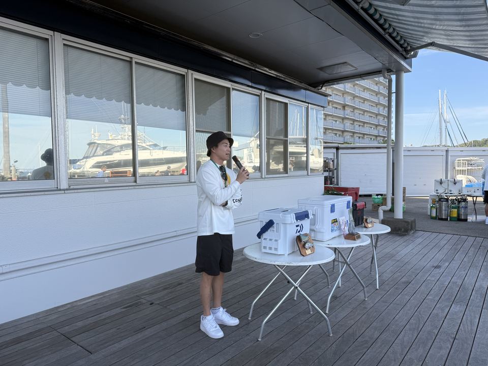 クラブハウス前のデッキでマイクを握る BOL全国幹事長の杉本氏と、賞品が並ぶテーブル