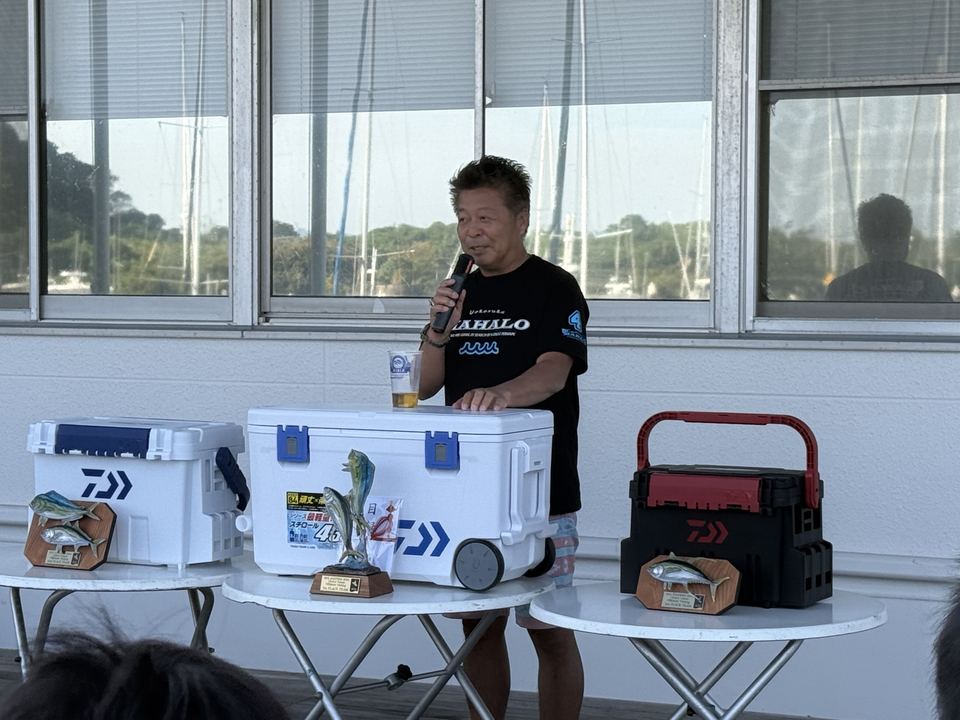 トロフィーと賞品が並ぶテーブルの後ろでマイクを握る MAHALO の谷本キャプテン